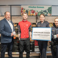 Four people standing in a grocery store near a fruit display, with two individuals in police uniforms holding a sign that reads, “Gemeinsam.sicher mit der REWE International AG.”