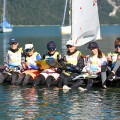 Six people in matching sailing gear sit on a wooden dock by a calm lake, each holding colorful books. Their feet dangle above the water. Sailboats and forested hills are visible in the background under a clear sky.
