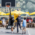 A group of people playing basketball on an outdoor court near a supermarket. A person in a police uniform is jumping to shoot the ball. Others are watching, with yellow umbrellas and fruit displays in the background.
