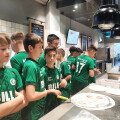 A group of young boys in green sports jerseys standing around a counter preparing pizzas in a kitchen. The kitchen has hanging black lights and tiled walls.