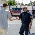 A young man in a gray and yellow outfit shakes hands with a police officer on a sunny outdoor basketball court. The officer wears a navy uniform and sunglasses. Other people and a police vehicle are visible in the background.