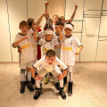 A group of young boys in identical white soccer uniforms posing indoors, with one boy wearing a festive Santa hat. Some are kneeling in front, others standing behind, all smiling or pointing playfully.
