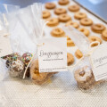 Bags of cookies with tags sit on a countertop, each containing treats, including round cookies with sprinkles and powdered sugar. A tray of similar cookies is in the background.