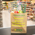 A paper grocery bag labeled "Land aufs Herz: Gewalt gegen Frauen ist untragbar" filled with vegetables is placed on a checkout counter. The background shows the interior of a supermarket with shelves lined with various products.