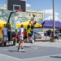 A group of young men playing a street basketball game on an outdoor court. One player is leaping toward the hoop with a basketball, while others watch. Yellow tents and a mural are visible in the background under a partly cloudy sky.