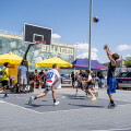Kids playing a game of street basketball on an outdoor court, surrounded by yellow umbrellas and a mural. One player is taking a jump shot. A police car is parked nearby under a clear blue sky.