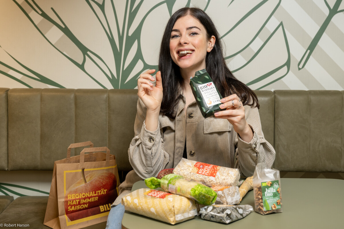 A person sits on a green bench, holding a package of organic nuts, and is smiling. In front of them are various snacks, including rolls, a carton of juice, and a paper bag with German writing. The background features a large leaf design on the wall.