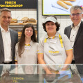 Four people, including two in bakery uniforms, stand smiling in front of a bakery display with fresh bread. Two men in suits are on either side of two bakery employees, a young woman and a young man, posing together. Bread and bakery signage are visible in the background.