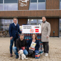 Four people are standing in front of a building, posing with a sign that reads "Feuerwerk, nein danke." One person is holding a small white dog on a leash. They are surrounded by packages of pet food.