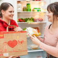 A woman in a red uniform hands a paper bag and food item to another woman in front of an open refrigerator filled with groceries. The bag features drawings of two children and a heart with "Danke!" written.