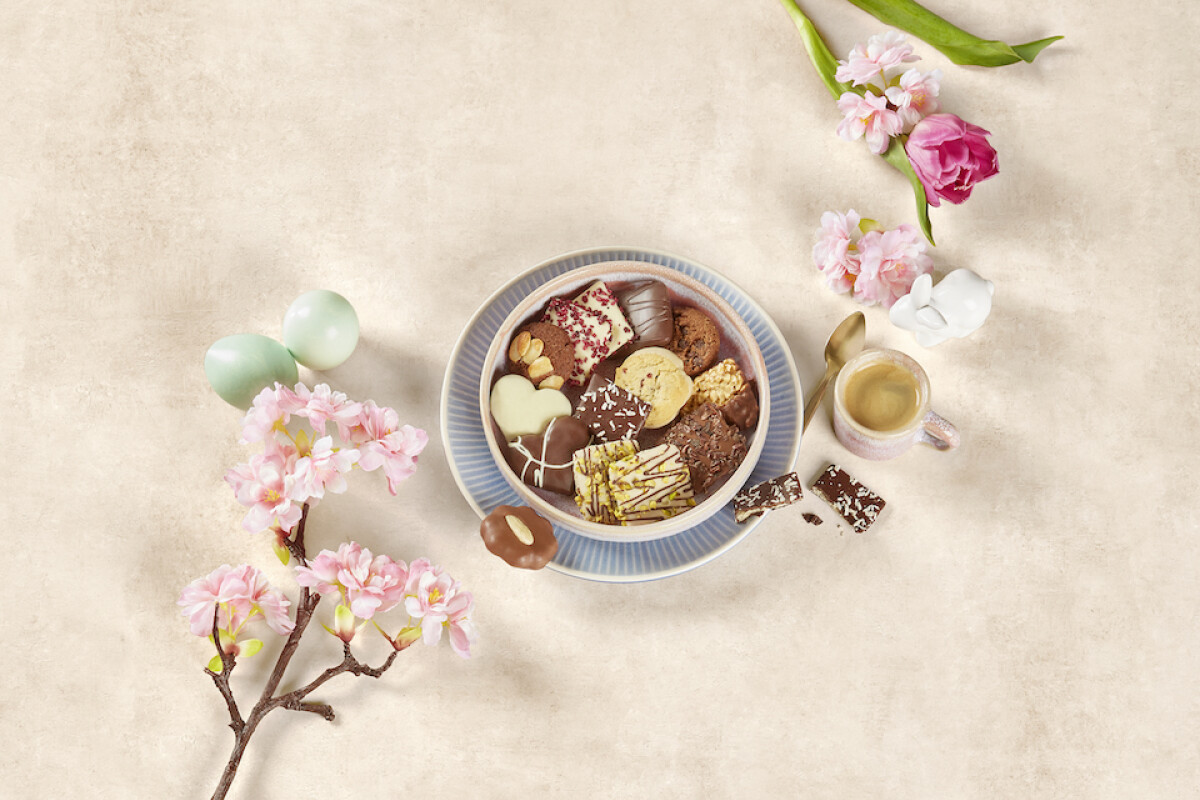 A bowl of assorted chocolates and cookies placed on a blue plate, accompanied by a small cup of espresso. Decorative elements include pink and white flowers, light blue eggs, and cherry blossom branches on a beige background.