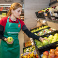 A grocery store employee wearing a green apron selects green apples from a display. The store has various fruits, including apples and melons, neatly arranged.