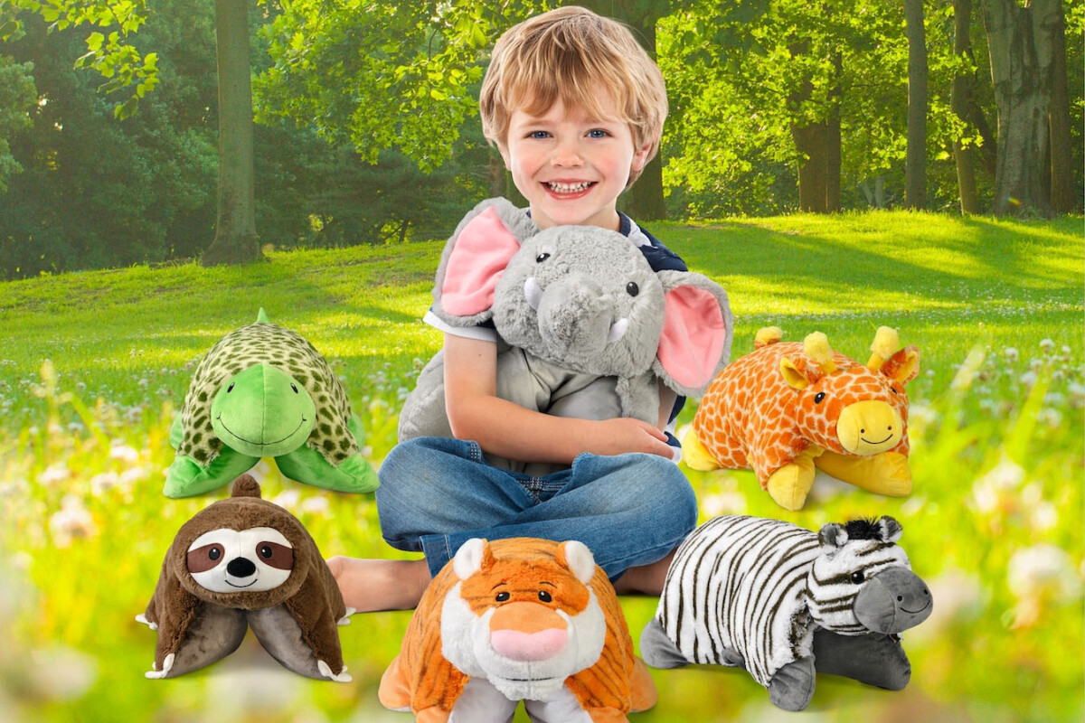 A child sitting on grass in a park, smiling and holding a plush elephant. Surrounding him are plush toys of a turtle, giraffe, sloth, tiger, and zebra. Sunlight filters through the trees in the background.