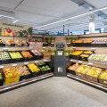 A supermarket produce section featuring neatly arranged fruits and vegetables, including bananas, oranges, apples, and pears. Shelves are labeled, and a small recycling station is visible in the center. Fluorescent lighting and modern fixtures illuminate the area.