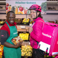 A grocery store employee and a food delivery worker smiling and posing in a produce section. The employee holds a bag of apples, while the delivery worker wears a pink helmet and carries a pink insulated delivery bag labeled "foodora." The background displays various fruits and vegetables with a sign reading "Frischer Genuss."