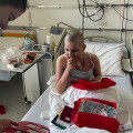 A person with a shaved head sits in a hospital bed, looking at a red gift bag. Another person stands nearby holding a photo frame. The room is equipped with medical devices.