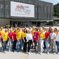 A group of people standing outside a building labeled "Skills Factory" in Schladming. They are wearing matching pink and yellow shirts, smiling, and posing for a group photo. The background includes a modern building and a mountainous landscape.