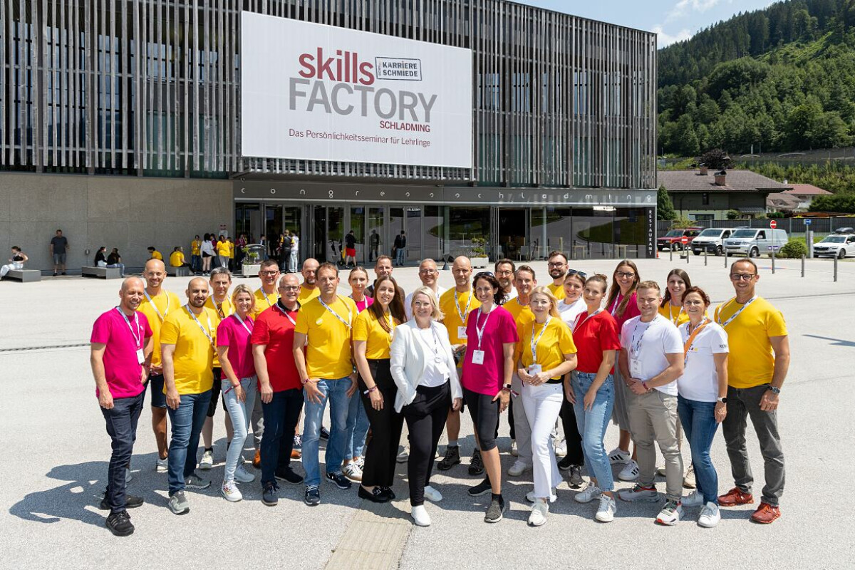 A group of people standing outside a building labeled "Skills Factory" in Schladming. They are wearing matching pink and yellow shirts, smiling, and posing for a group photo. The background includes a modern building and a mountainous landscape.