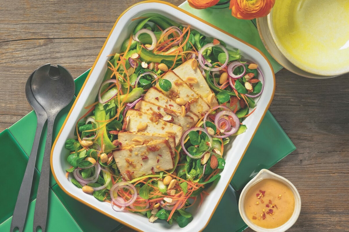 A colorful salad in a rectangular dish with grilled cheese slices, mixed greens, and sliced red onions, garnished with peanuts and radish spirals. Accompanying the dish are a set of salad servers, a small jug of dressing, and yellow flowers on a wooden table.
