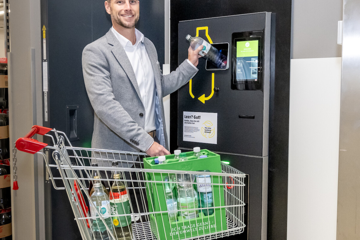 A man standing beside a bottle return machine, holding a glass bottle. He is wearing a light grey blazer and smiling. In front of him is a shopping cart filled with empty bottles. The wall sign above reads "Leergut Rückgabe."