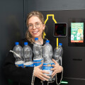 A person smiling and holding several empty plastic bottles in front of a recycling machine. They are wearing glasses and a black jacket with white trim.