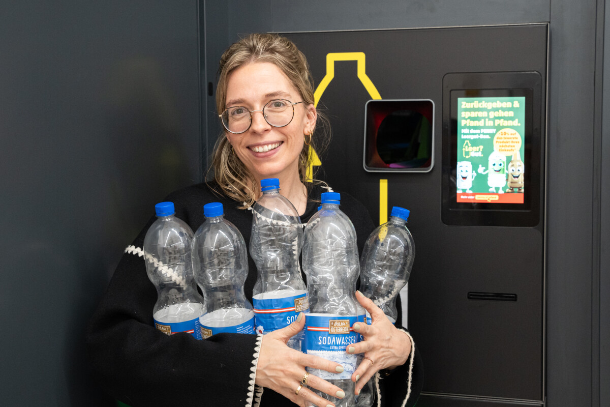 A person smiling and holding several empty plastic bottles in front of a recycling machine. They are wearing glasses and a black jacket with white trim.