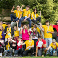 A large group of young people wearing yellow and pink shirts are gathered outdoors, posing playfully. Some are sitting and standing on a tree sculpture, while others are on the grass. They appear to be in a park with trees in the background. Everyone looks cheerful and energetic, with some making peace signs or waving. They are wearing name tags.