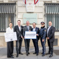 A group of six people, including a police officer and a person in a suit holding a sign that reads "GEMEINSAMSICHER – Eigener Dienst in Frauen," stand in front of a building labeled "Bundesministerium Inneres." Two other people hold a large plaque with text. Flags hang above the entrance.