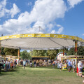 A lively outdoor festival with people walking around booths under a large, suspended canopy. The sky is blue with scattered clouds, and trees border the grassy area.