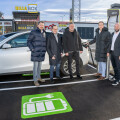 Five men standing next to an electric car at a charging station in a parking lot. The station is in front of a store with signs that read "BILLA BOX." A green electric vehicle charging symbol is painted on the ground.