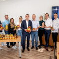 A group of people standing in an office, smiling and holding various jars of food products. A table in front of them displays more jars and food items. A banner with the logo "frey" is in the background.