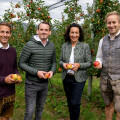 Four people standing in an orchard, each holding apples. They are smiling and dressed in a mix of casual and traditional clothing. Apple trees full of red apples are in the background.