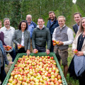 A group of people smiling around a large container filled with apples in an orchard. They are casually dressed and some are holding apples. The background shows rows of apple trees.