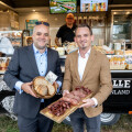 Two men in suits smiling and holding trays of various meats and bread in front of a food truck. The truck displays an assortment of pastries and desserts, with a man inside. The background shows trees and grassy area.