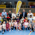 A group of children and adults posing together in a gymnasium. The children are dressed in casual sportswear, and two are holding certificates. Banners and logos are visible in the background.