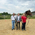 Three people standing in a sandy horse arena. A brown and white horse with a bridle is beside them. The person on the right is holding the horse's lead. A wooden fence, greenery, and a building are in the background under a cloudy sky.