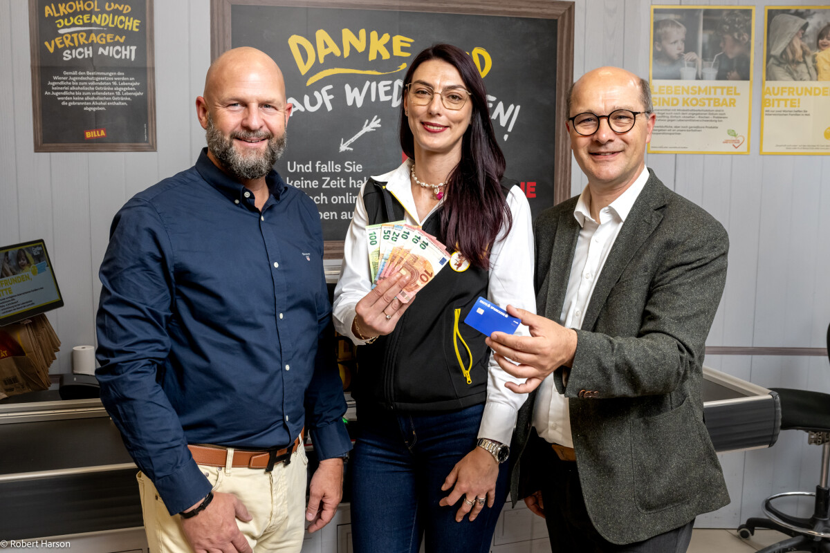 Three people stand in front of a chalkboard, smiling. The person in the middle holds a fan of euro banknotes, while the person on the right holds a blue card. The board has German text and the setting appears to be an indoor space.