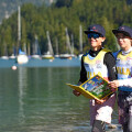 Two young sailors standing by a lake, wearing sailing vests and caps, holding a colorful book. In the background, sailboats are moored on the water with a forest and mountains in the distance.