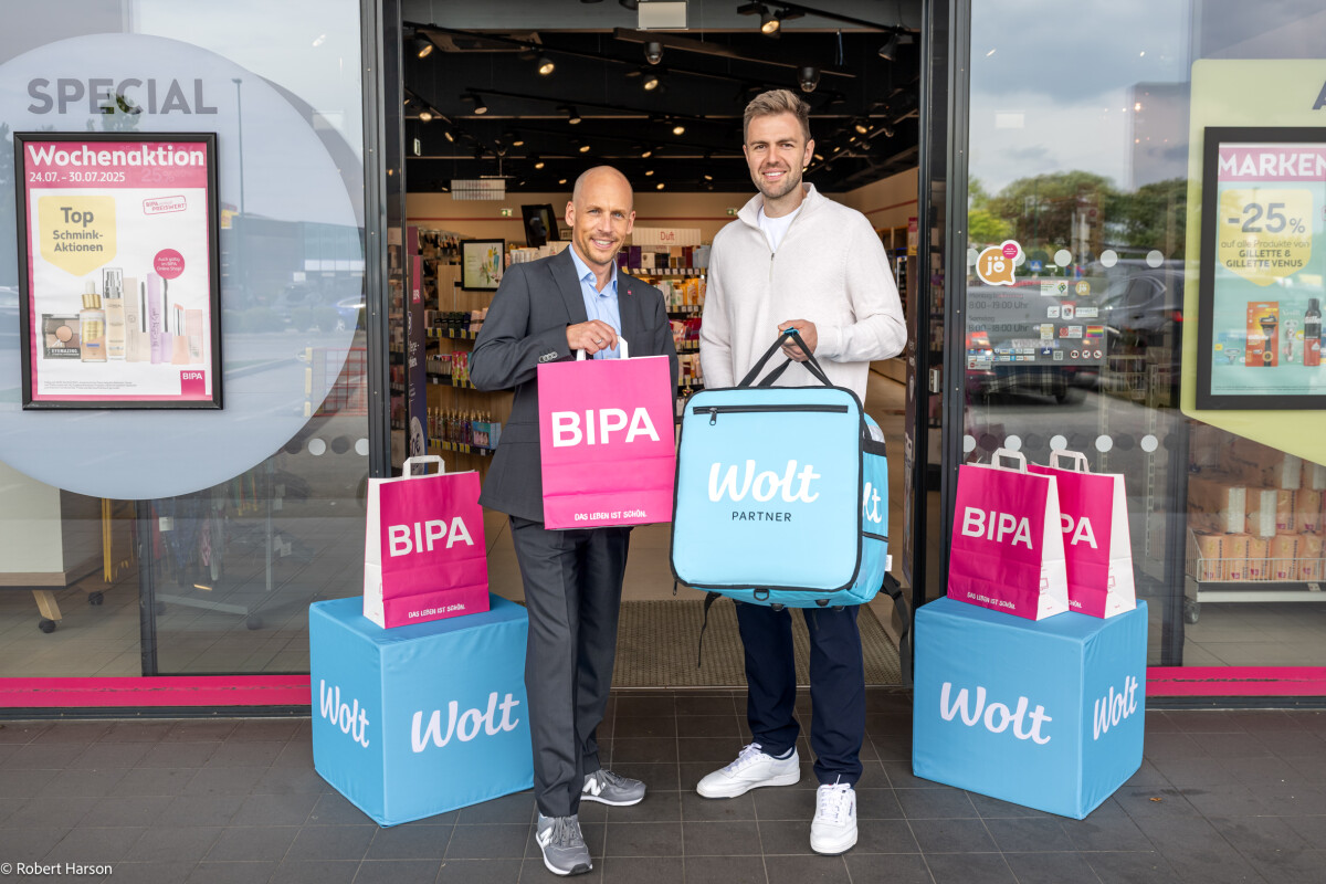 Two men smiling and holding delivery bags stand outside a store. One holds pink bags with "BIPA" on them, and the other holds a blue bag with "Wolt Partner." The store has glass doors and promotional posters.