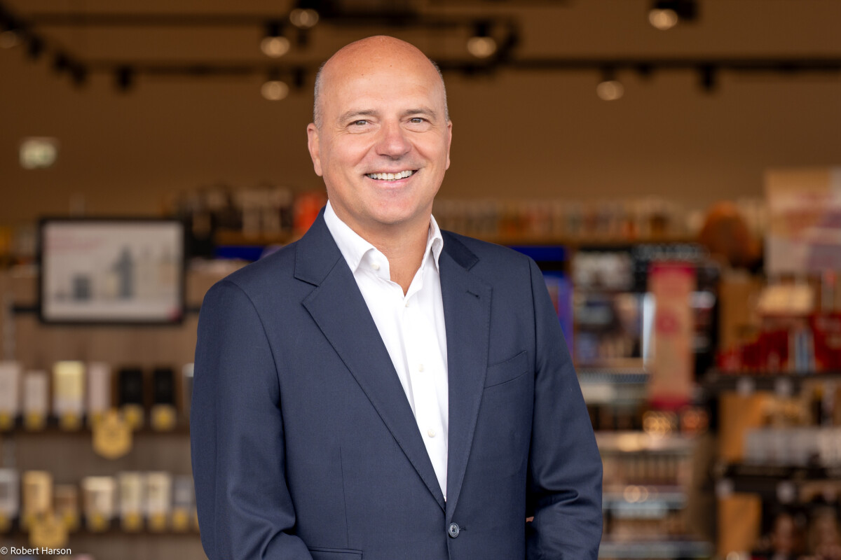 A bald man in a dark suit and white shirt smiling in a store setting with shelves and blurred products in the background.