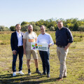 Four people stand in a grassy field under a clear sky, holding a sign that says "Blühendes Österreich" with the BILLA logo. They are smiling and dressed in casual and business attire. Trees are visible in the background.