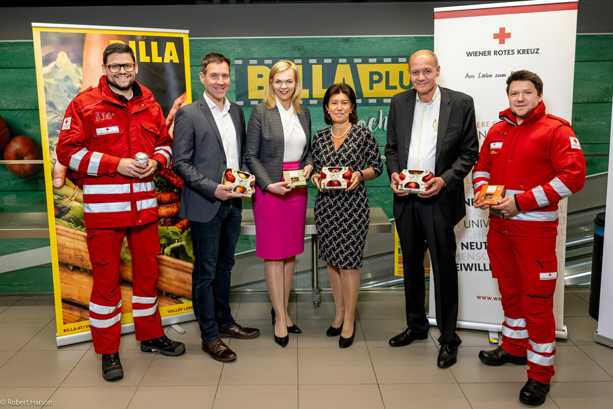 A group of six people standing in a row, holding packaged food items. Two individuals are wearing red uniforms with reflective stripes, likely from an emergency service. The others are dressed in business attire. Behind them are banners and a backdrop with the logo "BILLA PLUS" and another one with the Red Cross emblem. They are indoors on a tiled floor.