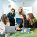 Five women gathered around a laptop in a modern, well-lit office space. They appear engaged and smiling, with plants and a sign in the background.
