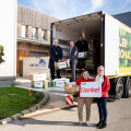 Four people unloading boxes from a truck, with two people in the foreground holding a sign that says "Danke!" A small tree and buildings are visible in the background.