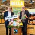 Two men standing in a grocery store near a display of organic products, including bananas and flowers. One man holds a "Fairbruary" sign, and the other holds a bouquet. Billa and Fairtrade logos are visible.