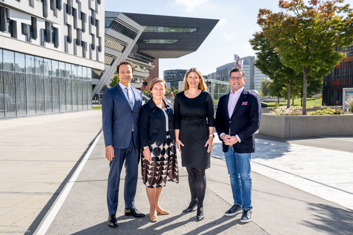 Four people are standing outside in a sunny urban setting. They are dressed in business attire, with modern buildings and a tree in the background. The sky is clear, and the environment appears bright and professional.