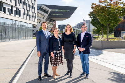 Four people are standing outside in a sunny urban setting. They are dressed in business attire, with modern buildings and a tree in the background. The sky is clear, and the environment appears bright and professional.
