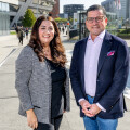 A man and a woman standing outside on a sunny day, smiling at the camera. They are both dressed in smart casual attire. The background features modern buildings, trees, and people walking.