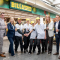 A group of people standing in front of a Billa Plus store, some in uniform, giving thumbs up and smiling. They are surrounded by store displays and pumpkins.
