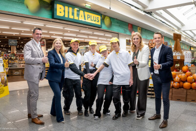 A group of people standing in front of a Billa Plus store, some in uniform, giving thumbs up and smiling. They are surrounded by store displays and pumpkins.